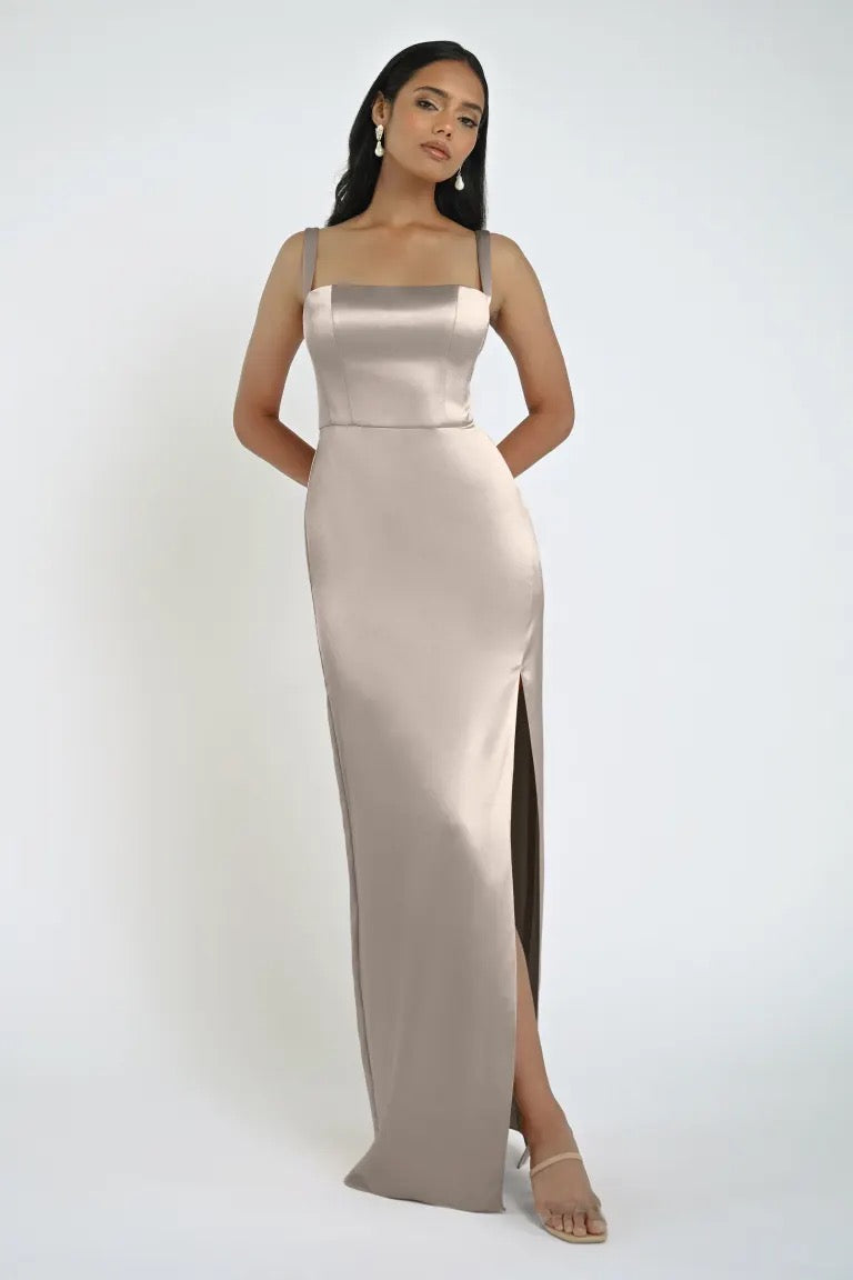 A woman wearing the Lowe Bridesmaid Dress by Jenny Yoo from Bergamot Bridal, a long and fitted satin gown with shoulder straps and a square neckline, poses against a plain white background. The sleek skirt features a side slit, and she completes the look with clear, strappy heels.