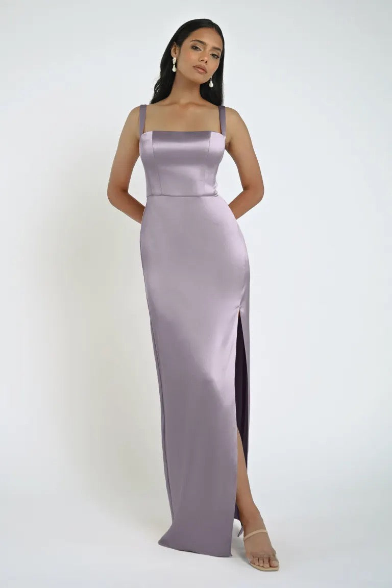 A woman in the floor-length, light purple satin Lowe Bridesmaid Dress by Jenny Yoo from Bergamot Bridal stands with her hands behind her back. She has long dark hair and wears dangling earrings and strappy heels, showcasing the dress's daring slit.