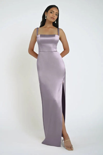 A woman in the floor-length, light purple satin Lowe Bridesmaid Dress by Jenny Yoo from Bergamot Bridal stands with her hands behind her back. She has long dark hair and wears dangling earrings and strappy heels, showcasing the dress's daring slit.