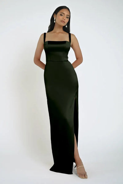 A woman stands against a plain white background, wearing the sleeveless black Lowe Bridesmaid Dress by Jenny Yoo from Bergamot Bridal, featuring a daring slit, and nude heels.