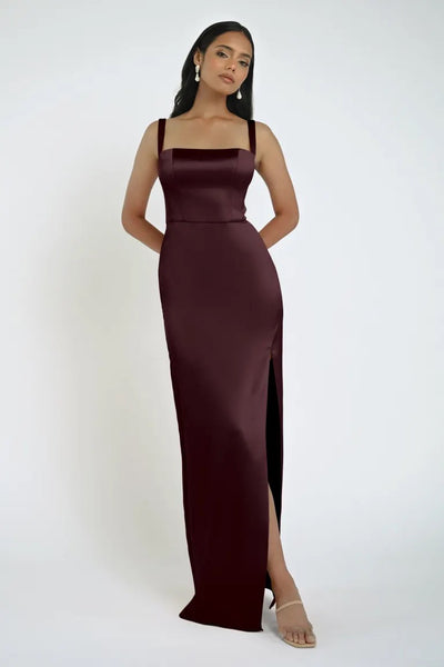 A woman in a sleeveless, floor-length Lowe Bridesmaid Dress by Jenny Yoo with a square neckline and side slit stands with her hands behind her back on a plain white background. This beautiful piece is offered by Bergamot Bridal.