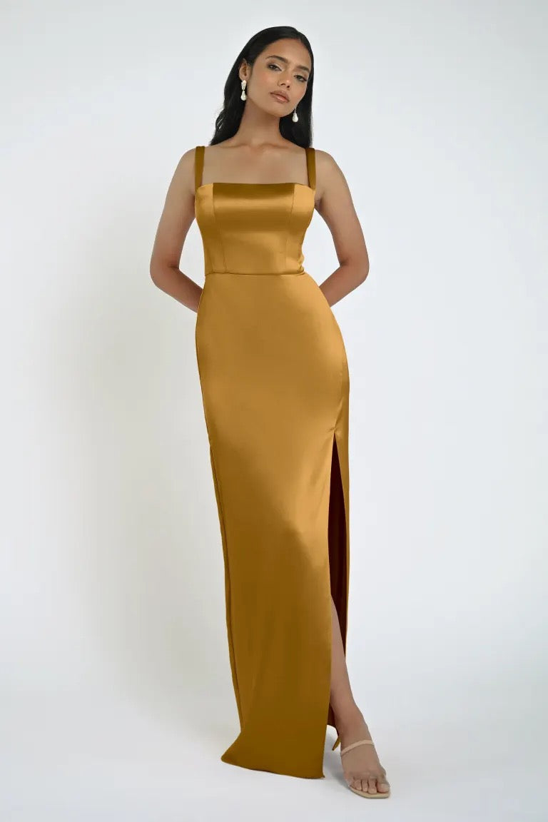 A woman stands against a plain white background wearing a sleek Lowe Bridesmaid Dress by Jenny Yoo, featuring a square neckline, sleeveless design, gold satin fabric, and a high slit on one side. She completes the look with tan sandals from Bergamot Bridal.