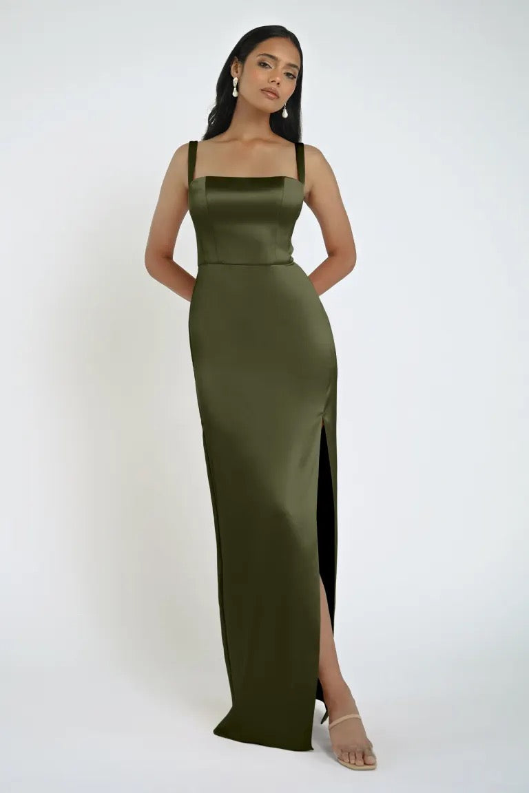 A woman is wearing a floor-length, olive green satin Lowe Bridesmaid Dress by Jenny Yoo from Bergamot Bridal. With a square neckline and a high slit, she stands with her hands behind her back, looking ahead.