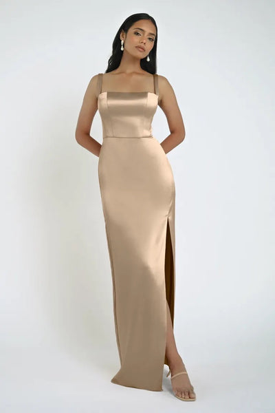 A woman stands facing the camera, adorned in a sleeveless Lowe Bridesmaid Dress by Jenny Yoo from Bergamot Bridal. The dress features a square neckline and a floor-length beige skirt with a side slit. She complements her look with dark hair, earrings, and heeled sandals.