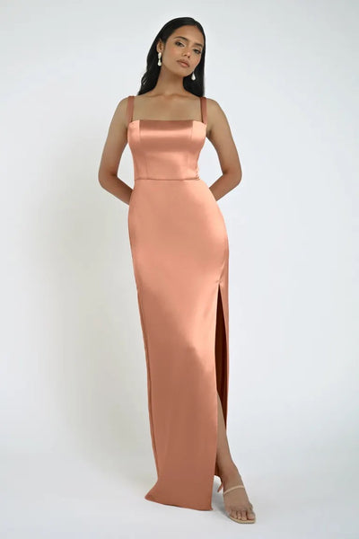 A woman in a sleeveless, peach-colored Lowe Bridesmaid Dress by Jenny Yoo, featuring a daring slit and square neckline, stands against a plain background, wearing simple earrings and open-toe heels.