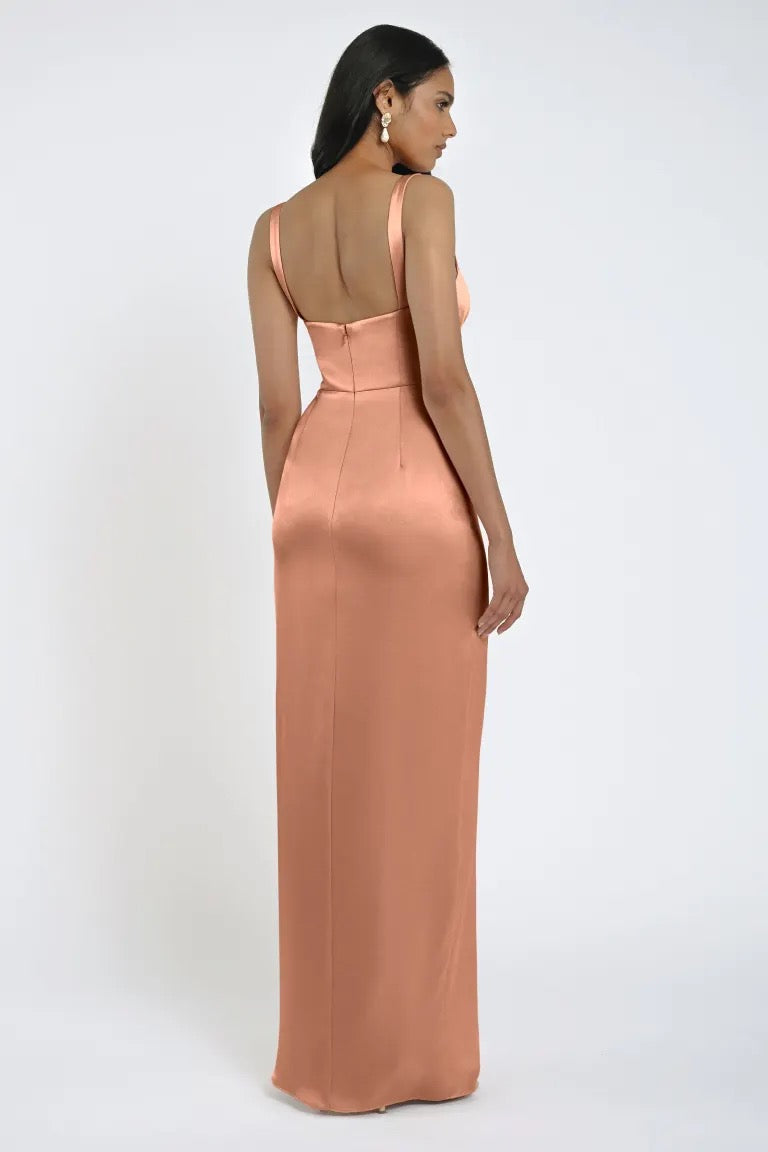 A woman in an elegant, shimmery, peach-colored Lowe Bridesmaid Dress by Jenny Yoo from Bergamot Bridal, featuring a square neckline, seen from the back with her hair down and wearing statement earrings.