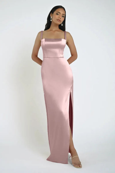 A woman stands posing in a stunning Bergamot Bridal Lowe Bridesmaid Dress by Jenny Yoo, featuring a sleek skirt and square neckline. The sleeveless, floor-length, pink satin ensemble boasts a thigh-high slit, complemented by her minimal jewelry and clear high-heeled sandals. The background is plain white.