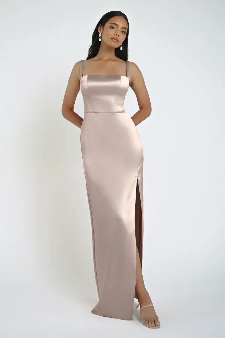 A woman stands wearing a long, silky Lowe Bridesmaid Dress by Jenny Yoo from Bergamot Bridal, featuring a square neckline and daring slit. She pairs it with nude heels and earrings.