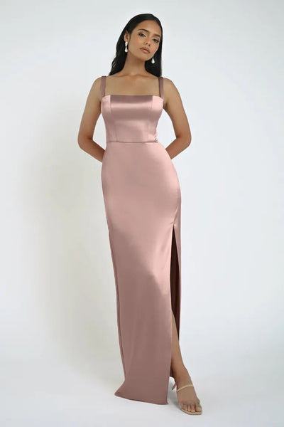 A woman is wearing a sleeveless, form-fitting, light pink satin Lowe Bridesmaid Dress by Jenny Yoo from Bergamot Bridal, featuring a high slit and square neckline. She is standing against a plain background.