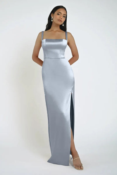 A woman stands against a plain backdrop wearing a stunning Lowe Bridesmaid Dress by Jenny Yoo, featuring a silver satin floor-length gown with thin straps, a daring slit, and a square neckline. She pairs the look with nude heels and gold earrings. This elegant ensemble is presented by Bergamot Bridal.