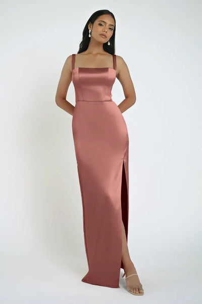 A woman in the Bergamot Bridal’s Lowe Bridesmaid Dress by Jenny Yoo, showcasing a sleeveless design with a square neckline and a sleek, long pink satin skirt with a high slit, stands against a plain white background.