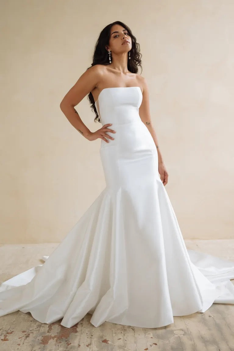 A woman in a strapless Lucia gown by Bergamot Bridal, featuring a fit and flare skirt, stands against a neutral background.