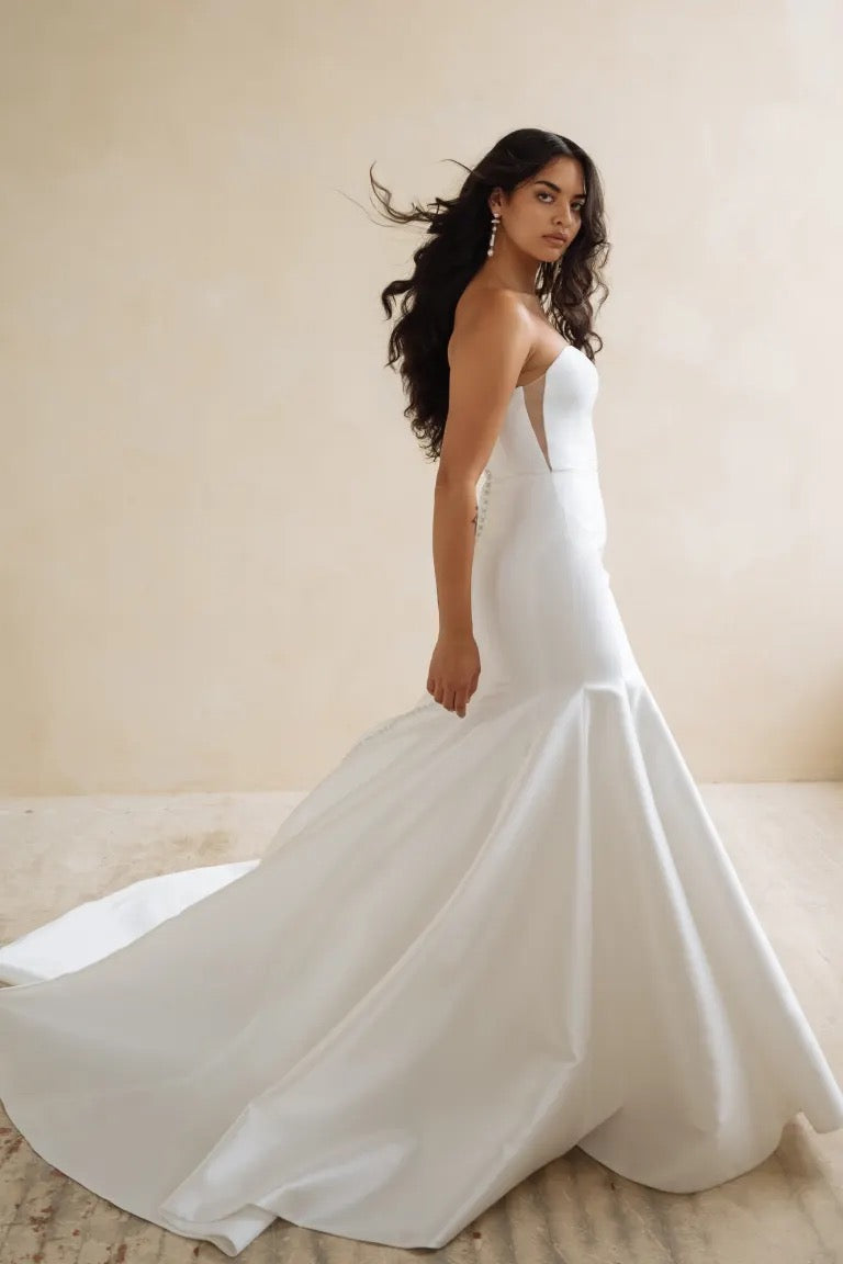 A woman gracefully poses against a neutral background in Bergamot Bridal's Lucia wedding dress by Jenny Yoo, showcasing the elegant silhouette of its fit and flare skirt and long train.