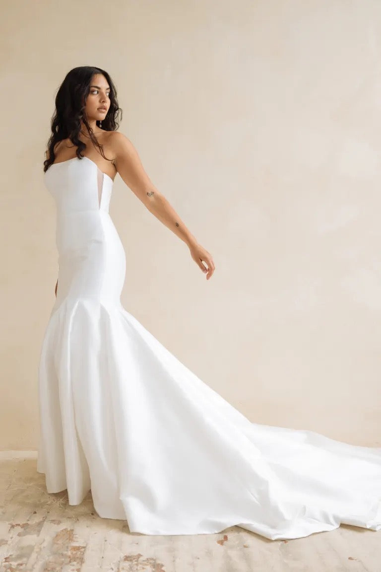 A woman elegantly poses in a neutral-toned room wearing the Lucia gown by Bergamot Bridal, a Jenny Yoo wedding dress with a strapless fit-and-flare design and an extended train.