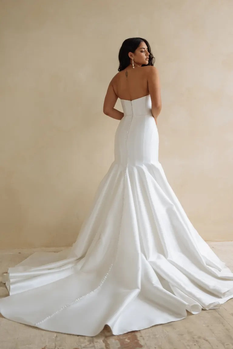 A woman in the Lucia wedding dress by Bergamot Bridal exudes elegance as she stands in a strapless gown made from canopy taffeta, with the fit and flare skirt sweeping into a long train against the neutral background, facing away.