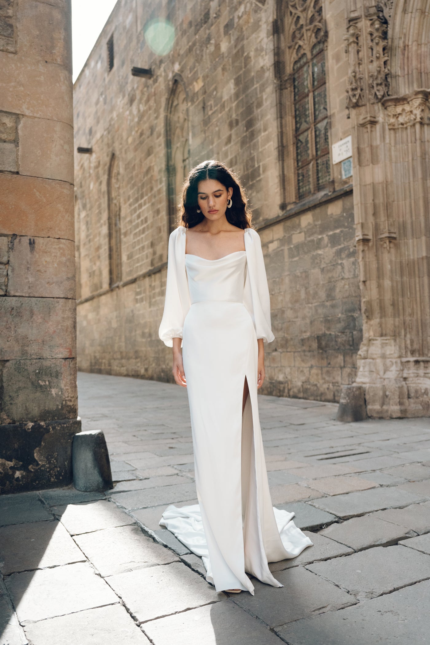 A woman in a Luciana - Jenny Yoo wedding dress by Bergamot Bridal, featuring oversized sleeves, stands on a cobblestone street near a historic stone building, with sunlight illuminating her from the back.