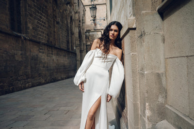 A woman wearing a white Luciana - Jenny Yoo Wedding Dress from Bergamot Bridal, featuring an off-shoulder design and oversized sleeves, leans against a stone wall in an old alley.