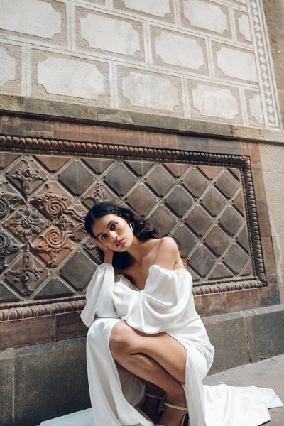 A woman in a Jenny Yoo wedding dress from Bergamot Bridal, featuring the Luciana design with oversized sleeves, sits against a patterned stone wall, looking at the camera.