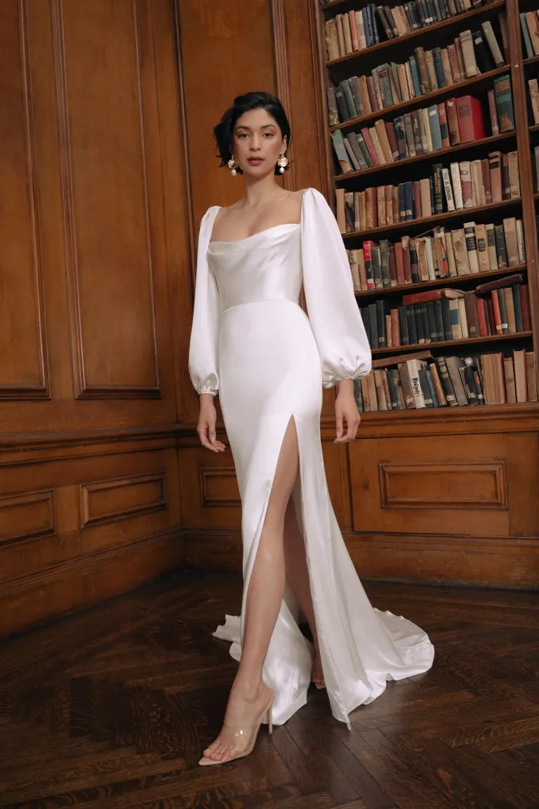 A woman in an elegant white Jenny Yoo Wedding Dress with a thigh-high slit poses against a backdrop of bookshelves from Bergamot Bridal.