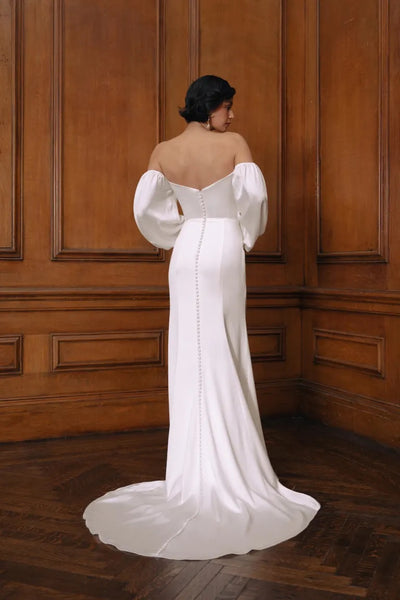 An elegantly dressed woman in a white Jenny Yoo Wedding Dress gown with a cowl neckline, standing with her back to the camera.