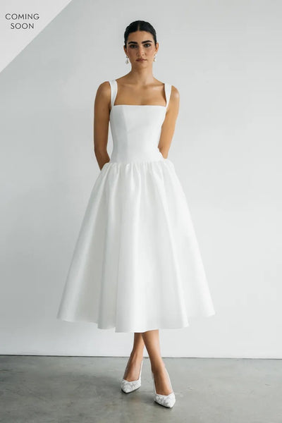 A person in the Lucille sleeveless, square-neck little white dress by Bergamot Bridal stands against a plain white wall, wearing white pointed heels. Text reads "Coming Soon.