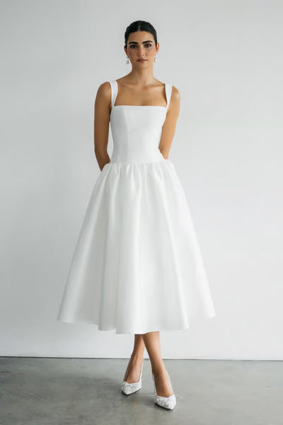 A woman poses against a plain backdrop in the Lucille Little White Dress by Bergamot Bridal—a tea-length, sleeveless taffeta dress with a fitted bodice and full skirt—styled elegantly with white pointed-toe heels.