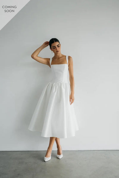 A woman in the elegant Lucille - Jenny Yoo Little White Dress from Bergamot Bridal poses against a plain white wall with one hand on her head.