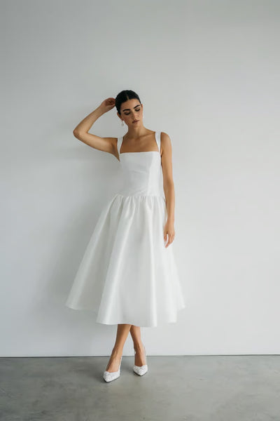 A woman wears the Lucille - Jenny Yoo Little White Dress by Bergamot Bridal with white heels, standing against a plain white wall, one hand touching her head and the other resting by her side.