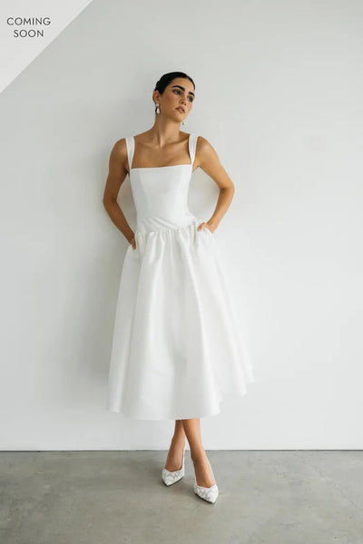 A person in a Lucille - Jenny Yoo Little White Dress by Bergamot Bridal stands against a white wall, looking to the side with hands in pockets. "Coming Soon" text graces the top left corner, highlighting the elegance of its structured bodice.