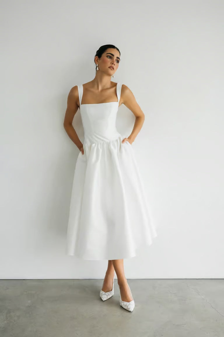 A woman wears the Lucille - Jenny Yoo Little White Dress by Bergamot Bridal, featuring a structured bodice and square neckline, standing against a white wall with hands in pockets and styled with white heels.
