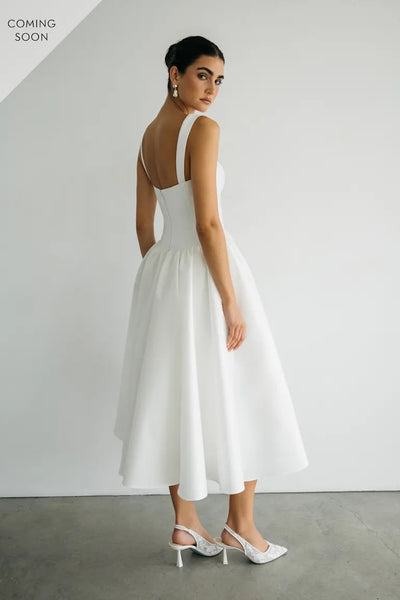 A woman in the elegant Lucille - Jenny Yoo Little White Dress from Bergamot Bridal and heels stands against a plain background, looking over her shoulder. Text in the top left corner reads "Coming Soon.