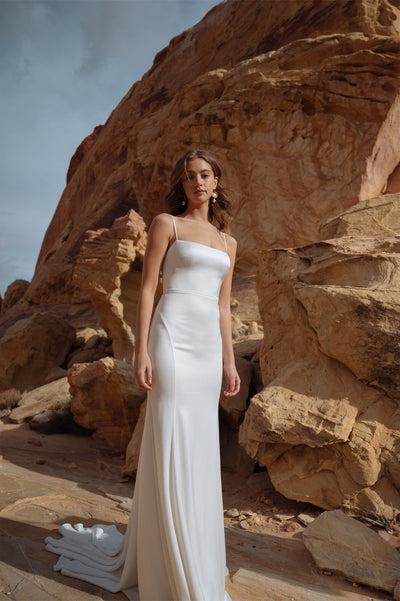 A woman in an elegant Lydia gown from Bergamot Bridal, with a flattering fit, standing against a rocky desert backdrop.