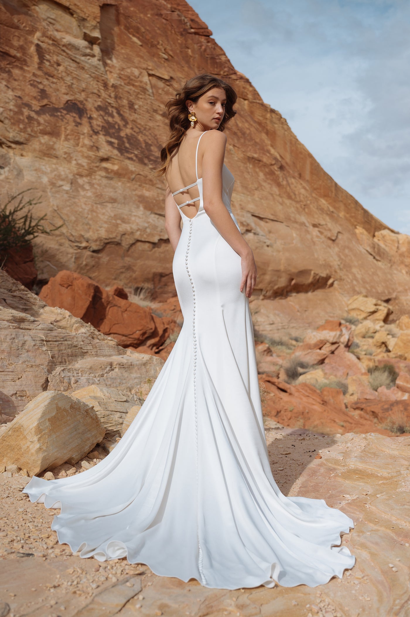 A woman in an elegant Jenny Yoo Wedding Dress with a trailing hem stands against a rocky desert backdrop.