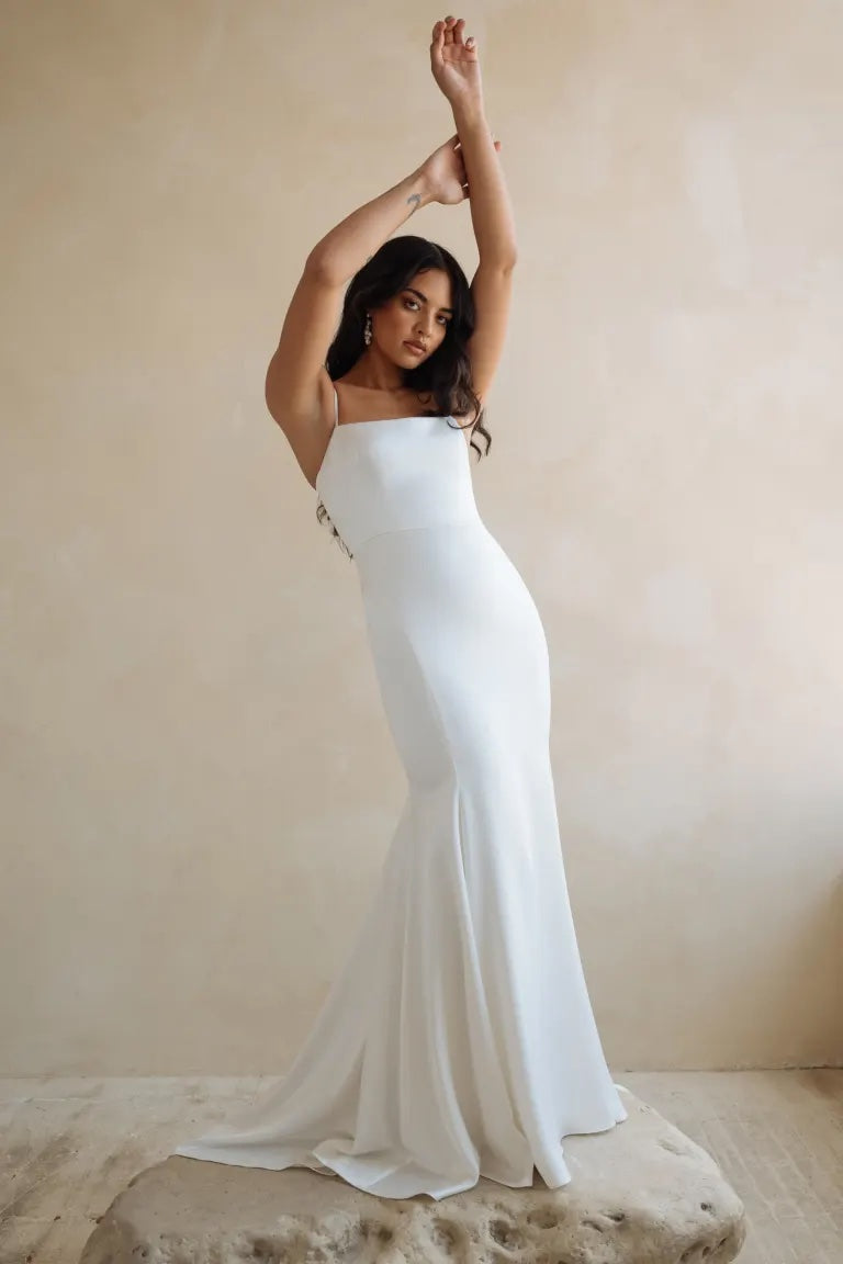 A woman in an elegant Jenny Yoo Wedding Dress slip gown from Bergamot Bridal posing with one arm raised.