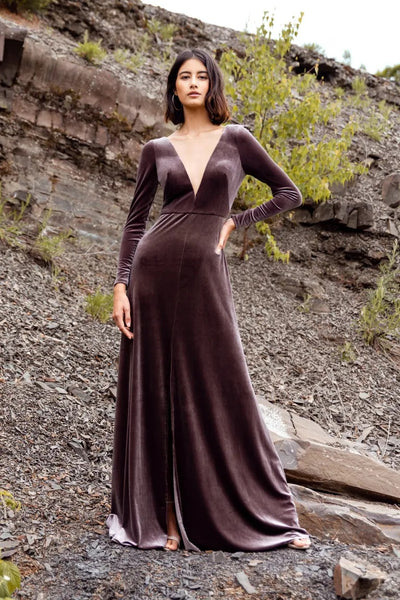 A woman in an elegant long-sleeve Malia - Bridesmaid Dress by Jenny Yoo gown standing against a rocky slope.