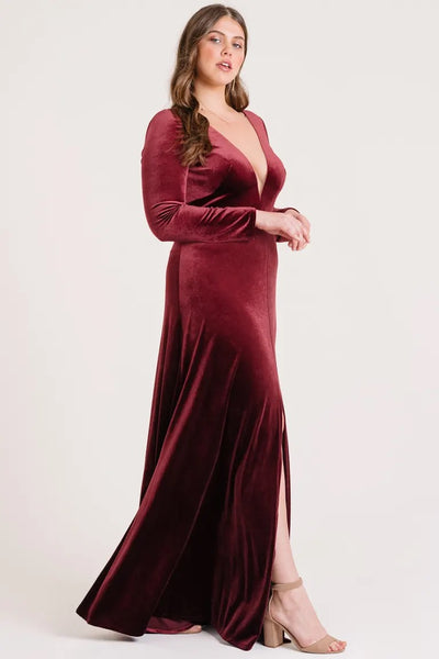 A woman posing in a long sleeve, deep red Malia - Bridesmaid Dress by Jenny Yoo gown with a front slit from Bergamot Bridal.
