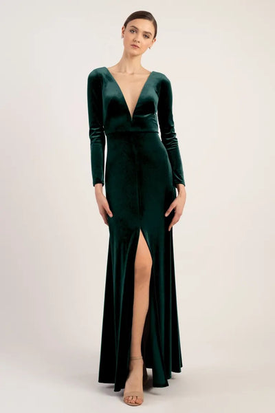 A woman in an elegant Luxe Velvet Malia bridesmaid dress by Jenny Yoo with a deep v-neckline and a thigh-high slit stands against a neutral background at Bergamot Bridal.