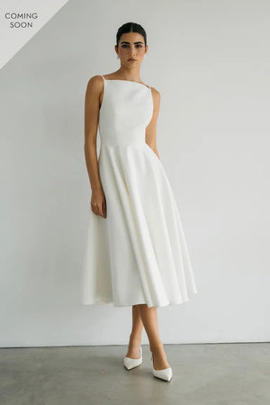 A woman models the Maria - Jenny Yoo Little White Dress by Bergamot Bridal, paired with white heels against a plain backdrop, radiating classic beauty with its romantic silhouette. "Coming Soon" text hints at what's next.