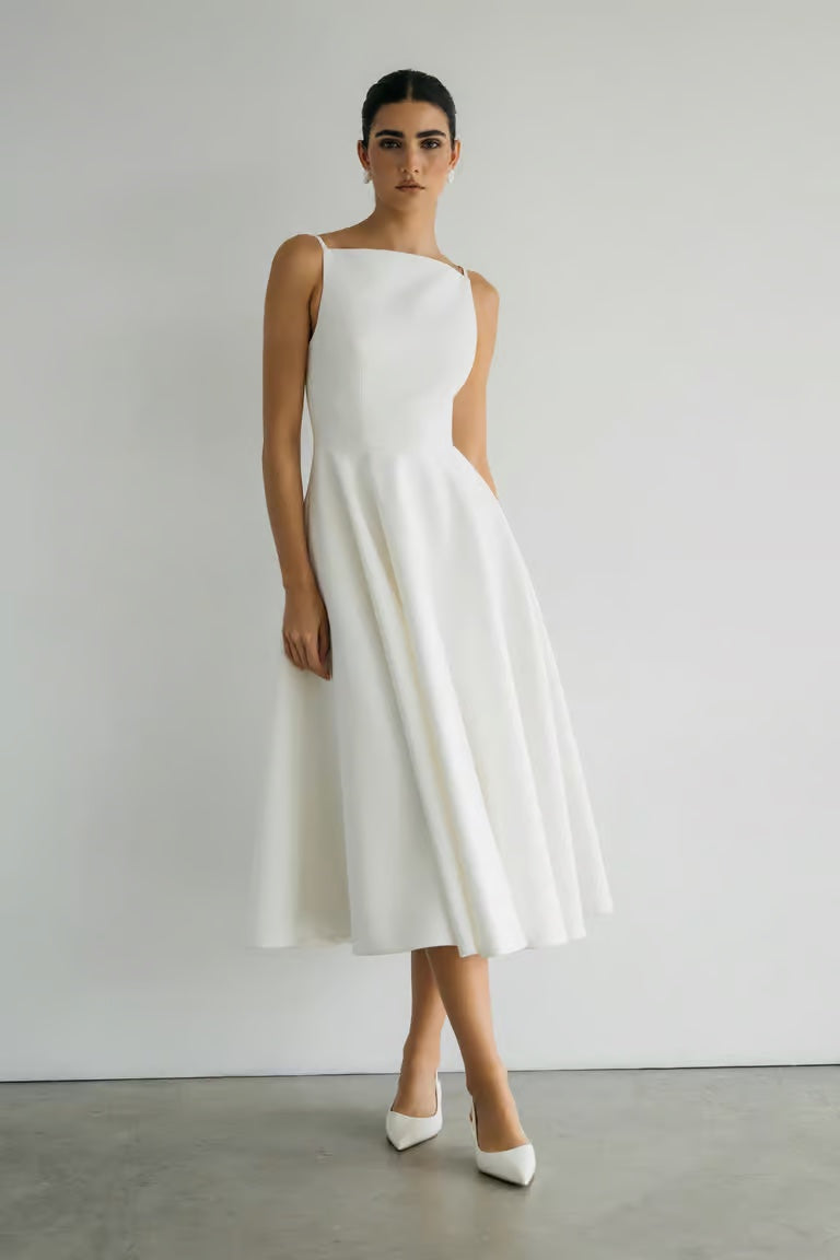 A woman stands against a white wall wearing the Maria - Jenny Yoo Little White Dress by Bergamot Bridal—sleeveless, mid-length, luxe faille fabric—paired with white pointed-toe heels for timeless style.