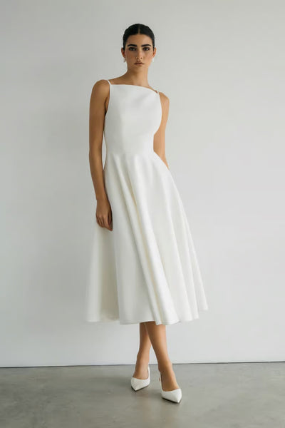 A woman stands against a white wall wearing the Maria - Jenny Yoo Little White Dress by Bergamot Bridal—sleeveless, mid-length, luxe faille fabric—paired with white pointed-toe heels for timeless style.