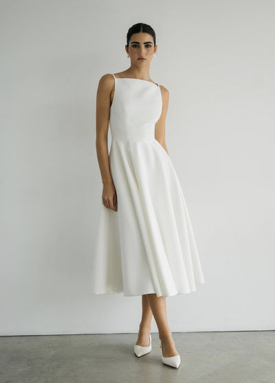 A woman in the Maria - Jenny Yoo Little White Dress by Bergamot Bridal, featuring a sleeveless, mid-length silhouette with a fitted bodice and flared skirt in luxe faille fabric, stands against a plain white wall.