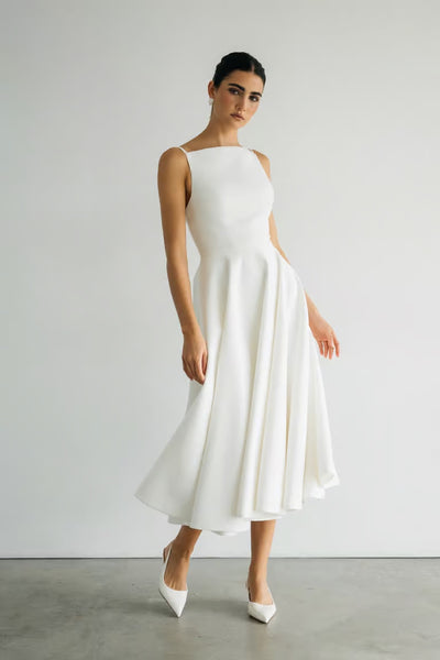 A woman stands against a plain white wall wearing the Maria - Jenny Yoo Little White Dress by Bergamot Bridal, a sleeveless, white midi dress with a flared skirt and romantic silhouette, paired with white pointed-toe heels.