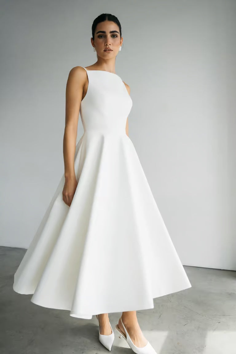 A woman stands indoors against a plain wall, wearing the Maria - Jenny Yoo Little White Dress by Bergamot Bridal—a sleeveless, white A-line midi dress with a boat neckline and white heels for an effortlessly timeless, romantic look.