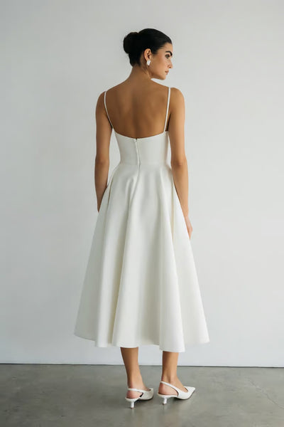 A woman faces away, wearing the Maria - Jenny Yoo Little White Dress by Bergamot Bridal, a white sleeveless midi dress in luxe faille with thin straps and a low back, paired with white heels. Her hair is in a bun for a classic look.