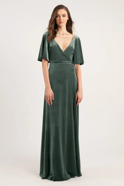 A woman modeling a dark green, v-neck wrap dress with an adjustable waist tie, against a neutral background from Bergamot Bridal's Marin - Bridesmaid Dress by Jenny Yoo.