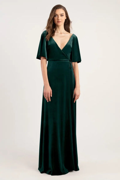 A woman models a Marin Bridesmaid Dress by Jenny Yoo in dark green velvet with a deep V-neck, short sleeves, and a true wrap design from Bergamot Bridal.