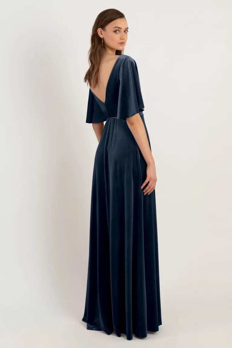 Woman in an elegant navy blue Marin - Bridesmaid Dress by Jenny Yoo with a v-back design and flutter sleeves posing over a neutral background from Bergamot Bridal.