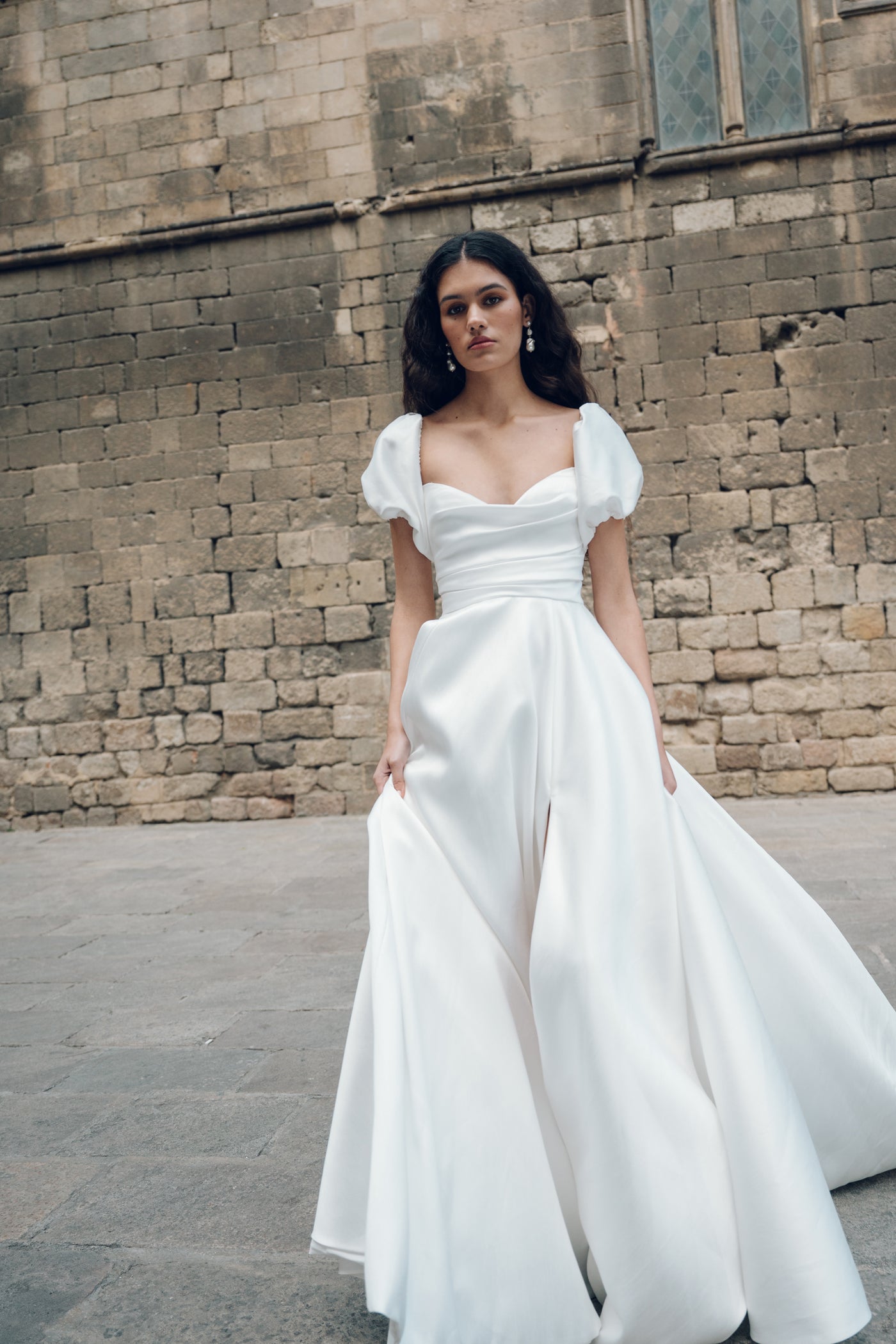 A woman in the Marisol wedding dress by Jenny Yoo from Bergamot Bridal, featuring a cowl neckline, stands gracefully before a textured stone wall.