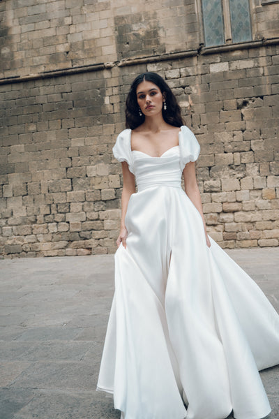 A woman in the Marisol wedding dress by Jenny Yoo from Bergamot Bridal, featuring a cowl neckline, stands gracefully before a textured stone wall.