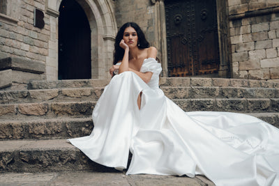 A woman in a floor-length Jenny Yoo wedding dress by Bergamot Bridal, featuring detachable puff sleeves, sits gracefully on stone steps before a historic building with wooden doors.